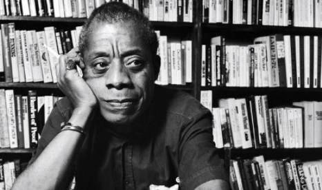 James Baldwin thinking In front of a packed bookshelf.