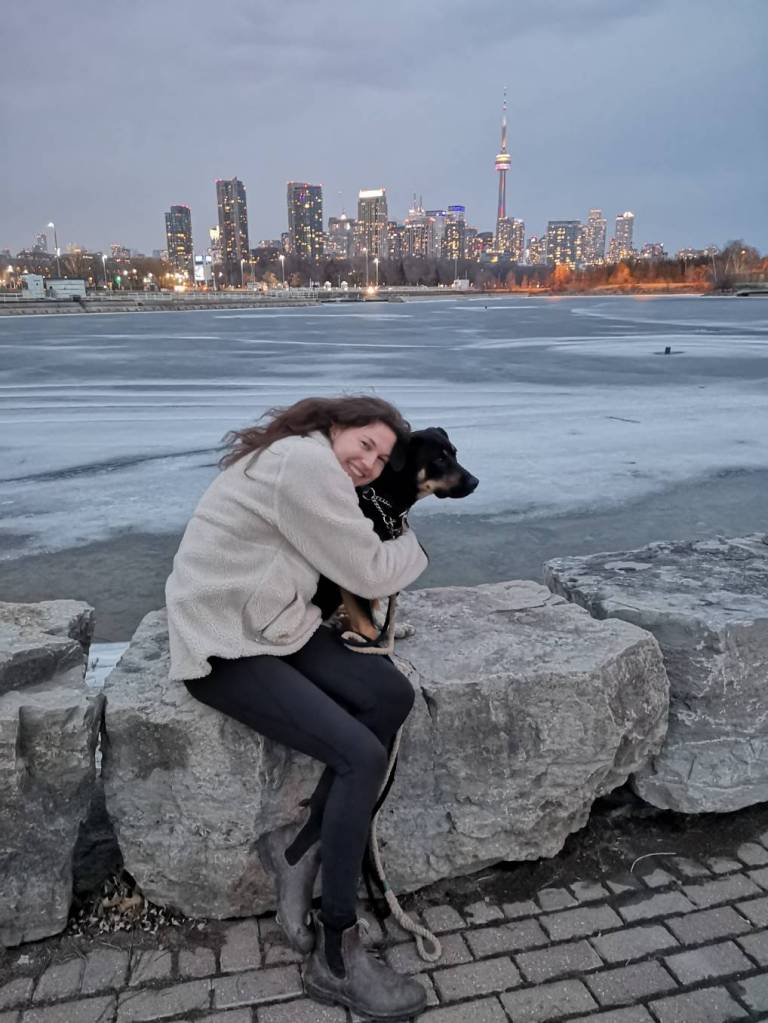 A girl hugging her dog in front of the Toronto city skyline at dusk.